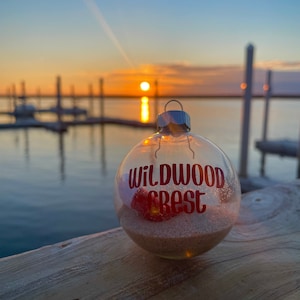 May include: A clear glass Christmas ornament with the words "Wildwood Crest" in red glitter lettering. The ornament is filled with sand and a red object. The background shows a sunset over the water.