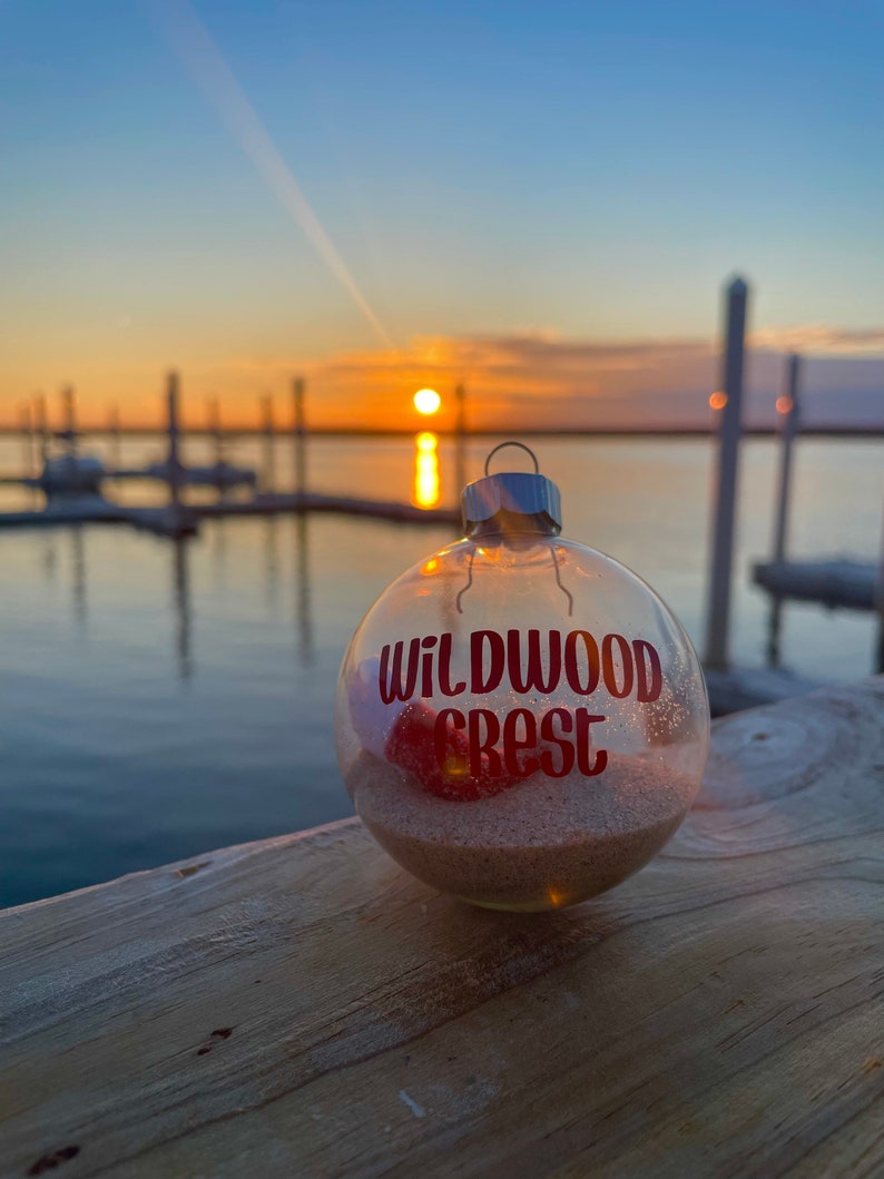 May include: A clear glass Christmas ornament with the words "Wildwood Crest" in red glitter lettering. The ornament is filled with sand and a red object. The background shows a sunset over the water.