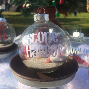 Puede incluir: Adorno de Navidad de vidrio transparente con una etiqueta blanca "stone harbor" y arena en su interior. El adorno est&aacute; sobre una base de metal marr&oacute;n.