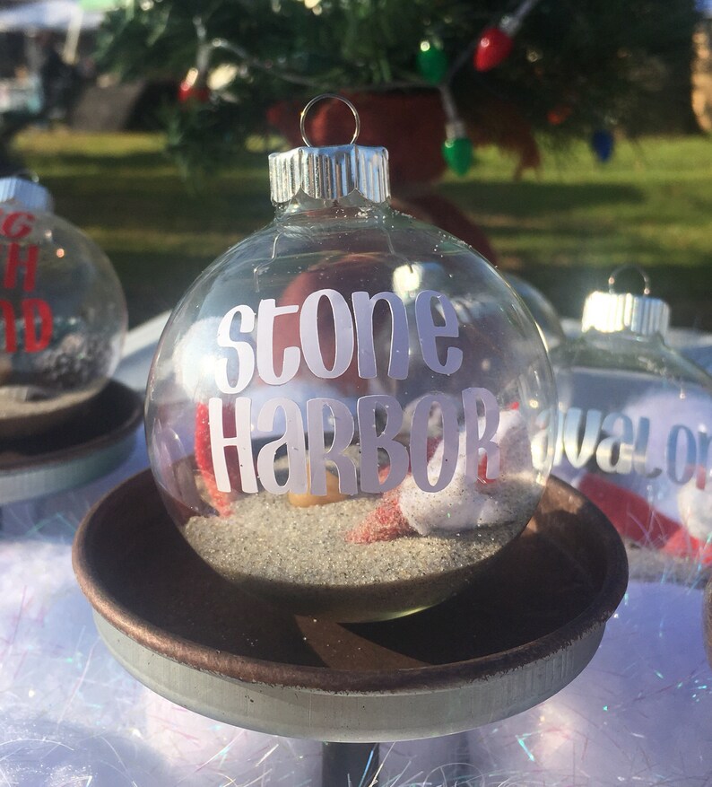 Puede incluir: Adorno de Navidad de vidrio transparente con una etiqueta blanca "stone harbor" y arena en su interior. El adorno est&aacute; sobre una base de metal marr&oacute;n.