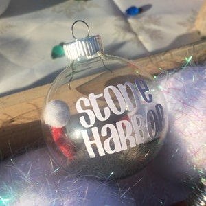 Puede incluir: Adorno navide&ntilde;o de vidrio transparente con un gorro de Pap&aacute; Noel de fieltro blanco y ribete rojo en el interior. El adorno tiene una parte superior plateada y una pegatina de vinilo blanco que dice "Stone Harbor".