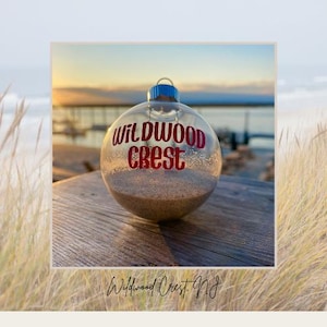 May include: Clear glass Christmas ornament filled with sand, featuring the text "Wildwood Crest" in red. The ornament has a blue cap and is set against a beach scene. The text "Wildwood Crest, NJ" is at the bottom.