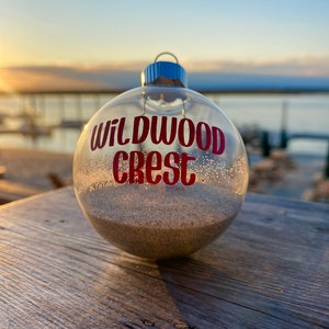 May include: Clear glass ornament filled with sand, featuring the text "WILDWOOD CREST" in red. The ornament has a silver cap with a blue loop. The background shows a sunset over a body of water.