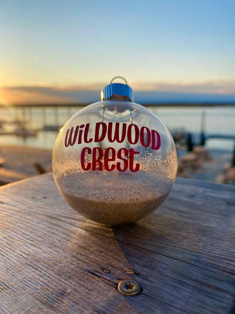 May include: Clear glass ornament filled with sand, featuring the text "WILDWOOD CREST" in red. The ornament has a silver cap with a blue loop. The background shows a sunset over a body of water.