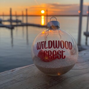 May include: Clear glass Christmas ornament with the words "Wildwood Crest" in red lettering. The ornament contains sand and a red seashell. The background shows a sunset over the water.