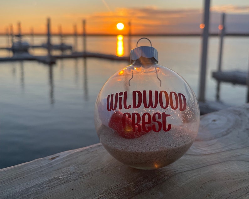 May include: Clear glass Christmas ornament with the words "Wildwood Crest" in red lettering. The ornament contains sand and a red seashell. The background shows a sunset over the water.