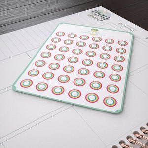May include: A sheet of 48 colourful circle stickers with a white background. The stickers have three concentric circles in pink, yellow, and blue. The sheet has a green border and the text "ALORA PLANNING DESIGNS" in the top centre.