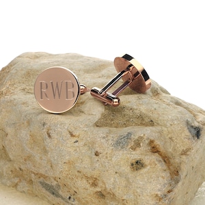 May include: Rose gold cufflinks with engraved initials "RWB" on a round face. The cufflinks are displayed on a textured, light-colored stone, showcasing their polished finish and design. A classic accessory.