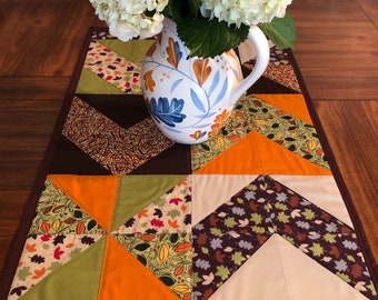 May include: A quilted table runner with a brown, orange, green, and white patchwork design featuring a leaf pattern. The runner has a brown border with a leaf pattern. A white pitcher with a blue and brown floral design is on top of the runner, filled with white hydrangeas.