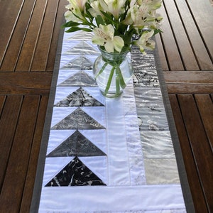 May include: A white table runner with a gray border and a pattern of black and white triangles. A vase with white flowers is on top of the runner.