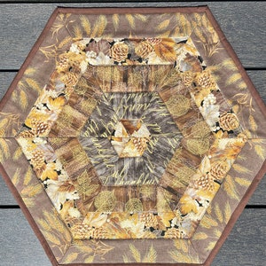 May include: A hexagonal table runner with a brown background and a geometric pattern of wheat, leaves, and pinecones in shades of gold and brown.