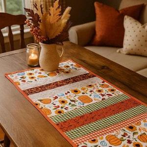 May include: A fall-themed table runner with a patchwork design featuring sunflowers, pumpkins, and fall leaves. The runner has a white background with orange, green, and brown accents. The runner is approximately 12 inches wide and 48 inches long.