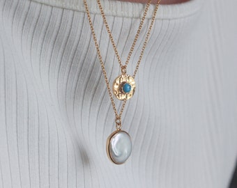 May include: A gold chain necklace with two pendants. The top pendant is a round, hammered gold disc with a turquoise stone in the center. The bottom pendant is a round, white pearl.