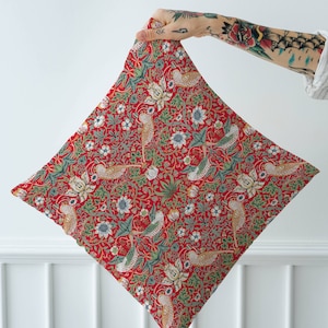 May include: Red fabric with a repeating pattern of stylized birds, flowers, and leaves. The fabric is folded in half and held by a hand.