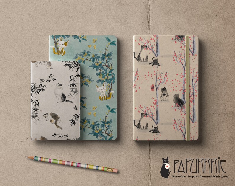 May include: Three different notebooks with cat illustrations. The notebooks have a variety of designs, including bamboo, flowers, and branches. The notebooks are all closed and have a white background. The text "PAPURRIE" is visible in the bottom right corner of the image.