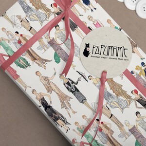 Op de afbeelding: Wit cadeaupapier met een herhalend patroon van vrouwen in jurken in de stijl van de jaren 1920. Het papier is vastgebonden met een roze lint en een rond, beige label met de tekst "PAPURRIE Perfect Paper - Created With Love".
