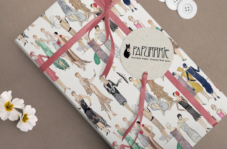 Op de afbeelding: Wit cadeaupapier met een herhalend patroon van vrouwen in jurken in de stijl van de jaren 1920. Het papier is vastgebonden met een roze lint en een rond, beige label met de tekst "PAPURRIE Perfect Paper - Created With Love".