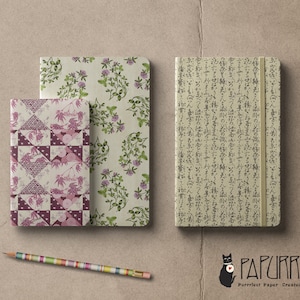 May include: Three different patterned notebooks, one with a floral design, one with a geometric pattern, and one with Japanese characters. The notebooks are on a brown surface with a pencil and a black cat logo with the text "Papurrie Purrfect Paper - Created With Love".