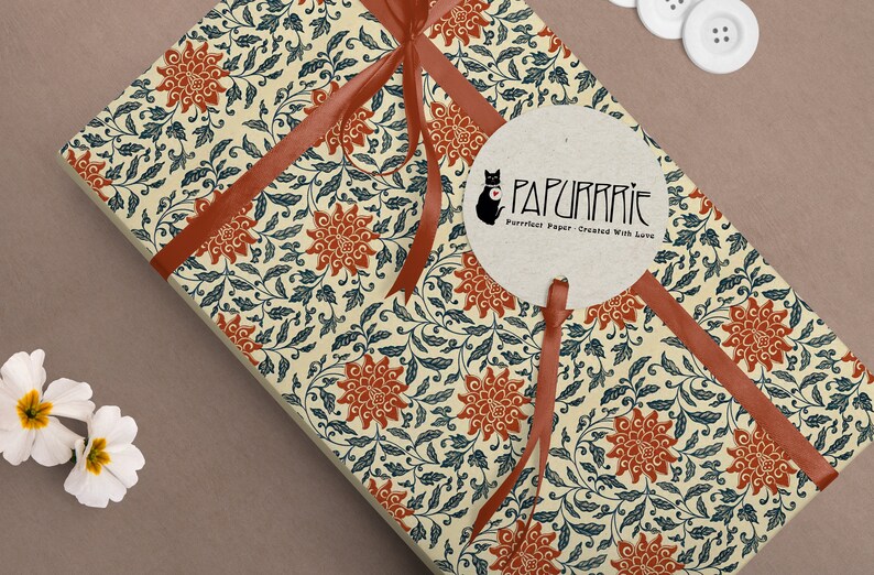 May include: A cream-colored gift wrap with a red ribbon and a repeating pattern of blue leaves and orange flowers. The gift wrap has a round sticker with a black cat and the text "PAPURRIE Perfect Paper - Created With Love".