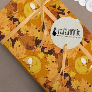 May include: A gift wrapped in orange and brown wrapping paper with a pattern of orange and brown leaves and sleeping orange cats. The paper is tied with a gold ribbon and a round brown paper tag with the text "PAPURRRIE" and "Purrfect Paper - Created With Love".