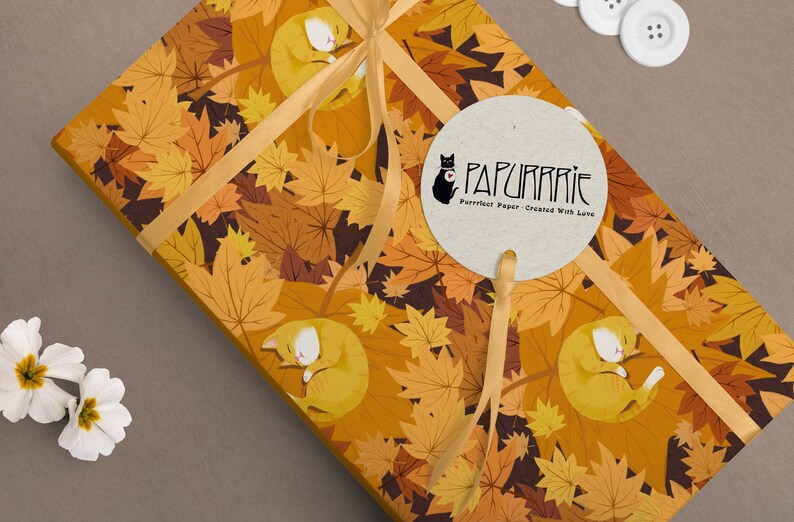May include: A gift wrapped in orange and brown wrapping paper with a pattern of orange and brown leaves and sleeping orange cats. The paper is tied with a gold ribbon and a round brown paper tag with the text "PAPURRRIE" and "Purrfect Paper - Created With Love".