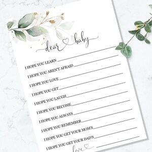以下が含まれることがあります： 「dear baby」という言葉がエレガントな筆記体で書かれた白いベビーシャワーゲームカード。ゲストが赤ちゃんへの願い事を書くための行があります。装飾的な緑と金のフローラルのアクセントが左上隅を飾っています。