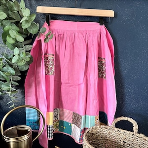 May include: A pink cotton apron with a patchwork bottom border and two floral print pockets. The apron is hanging on a wooden hanger against a dark blue background.