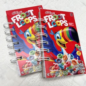 Puede incluir: Dos cuadernos con espiral hechos de cajas de cereales Kellogg's Froot Loops recicladas. Las cubiertas rojas presentan el logotipo de Froot Loops, una mascota tucán y cereales coloridos. Cada cuaderno está etiquetado "NET WT 27g".