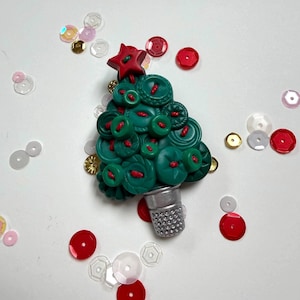 May include: A decorative Christmas tree ornament crafted from various shades of green buttons, featuring a red star at the top and a silver thimble as the base. The tree is accented with red thread stitching and surrounded by scattered red, white, and gold sequins.