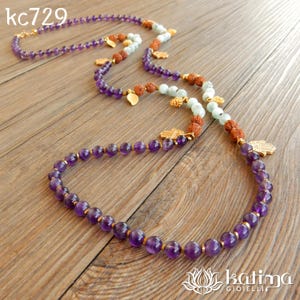 May include: A long necklace featuring purple amethyst beads, light green beads, and brown textured beads. The necklace includes gold-toned charms shaped like Hamsa hands and small leaves. The text kc729 is visible in the top left corner.