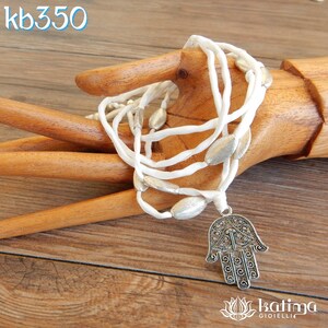 Può includere: Collana in cordoncino di seta bianco con perline ovali argentate e un ciondolo hamsa argentato. Il ciondolo hamsa è decorato con dettagli intricati ed è un simbolo di protezione.