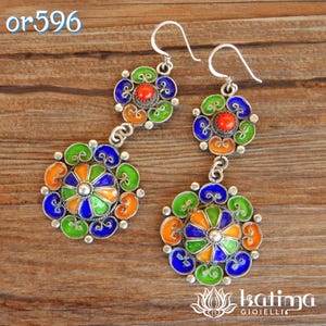 May include: A pair of silver earrings with three circular pendants each. The pendants are decorated with blue, green, orange, and red enamel and feature a small red gemstone in the center. The earrings are hanging from silver hooks.
