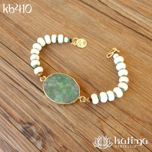 Può includere: Un braccialetto fatto a mano con una grande pietra verde ovale con macchie rosse, incastonata in una lunetta color oro. Il braccialetto è infilato con perline bianche e verdi, con una chiusura dorata e un piccolo ciondolo dorato. Il testo "kb410" è in alto a sinistra.