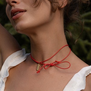 May include: A red choker necklace with a gold-coloured leaf pendant and a bow closure. The necklace is worn against a white top with ruffled edges. The background is a blurred green.