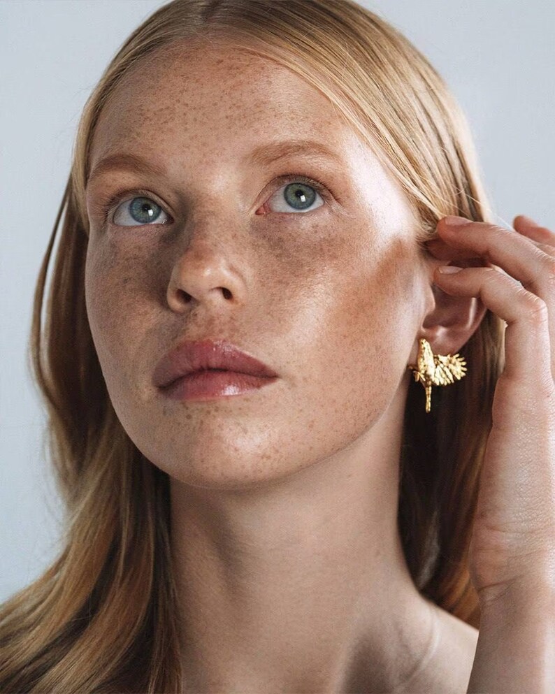 May include: A close-up of a woman's face with freckles. She is wearing a gold earring with a bird design.