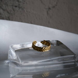 May include: A gold-colored ring with a leaf design sits on a clear glass block. The ring is a simple band with a delicate leaf pattern.