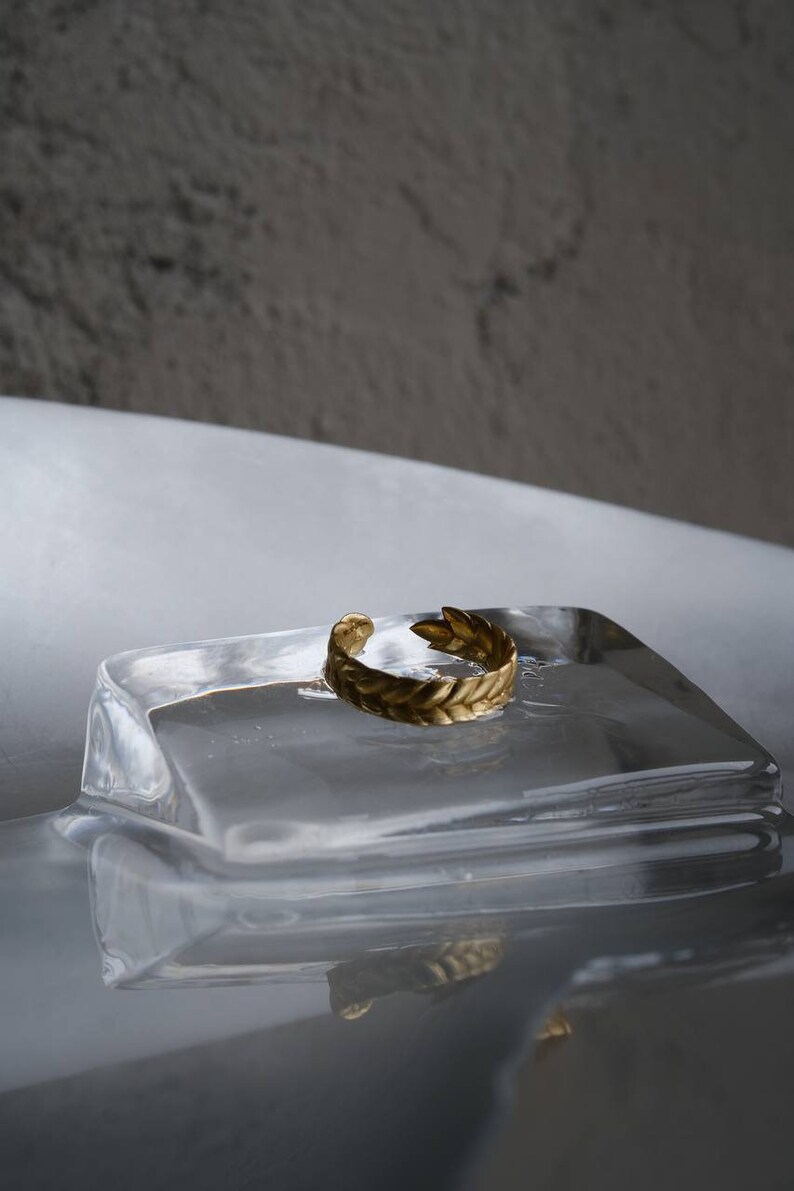 May include: A gold-colored ring with a leaf design sits on a clear glass block. The ring is a simple band with a delicate leaf pattern.