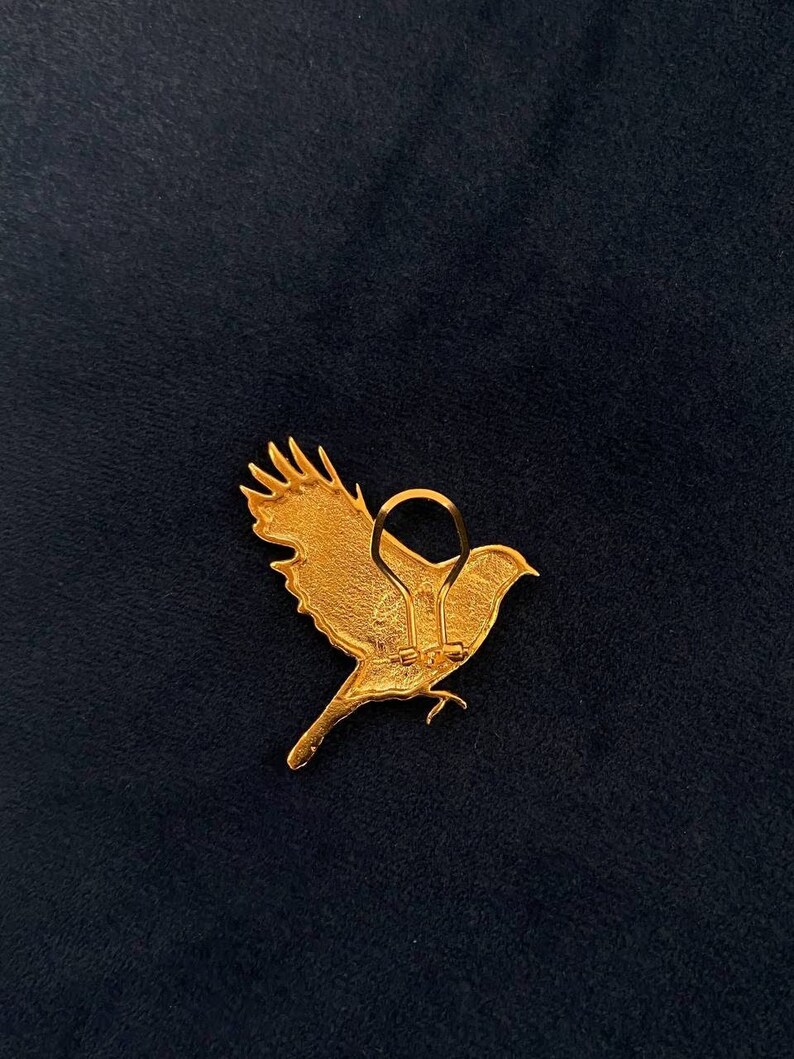 May include: Gold-coloured metal bird brooch with a detailed design. The brooch has a hinged clasp on the back.