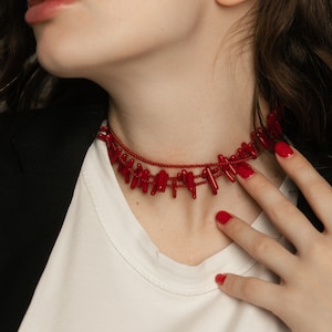 May include: A woman wearing a white shirt and a black blazer with a red coral choker necklace. Her nails are painted red.