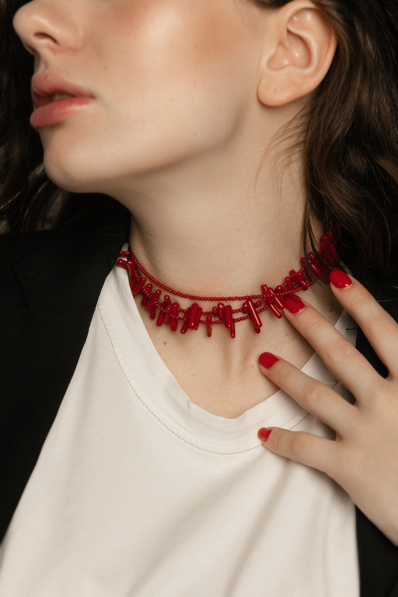 May include: A woman wearing a white shirt and a black blazer with a red coral choker necklace. Her nails are painted red.