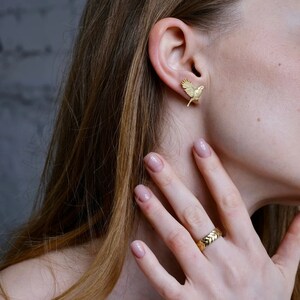May include: A gold bird-shaped earring with a textured surface. The earring is worn in the left ear of a person with long brown hair. The person is also wearing a gold ring on their right ring finger.