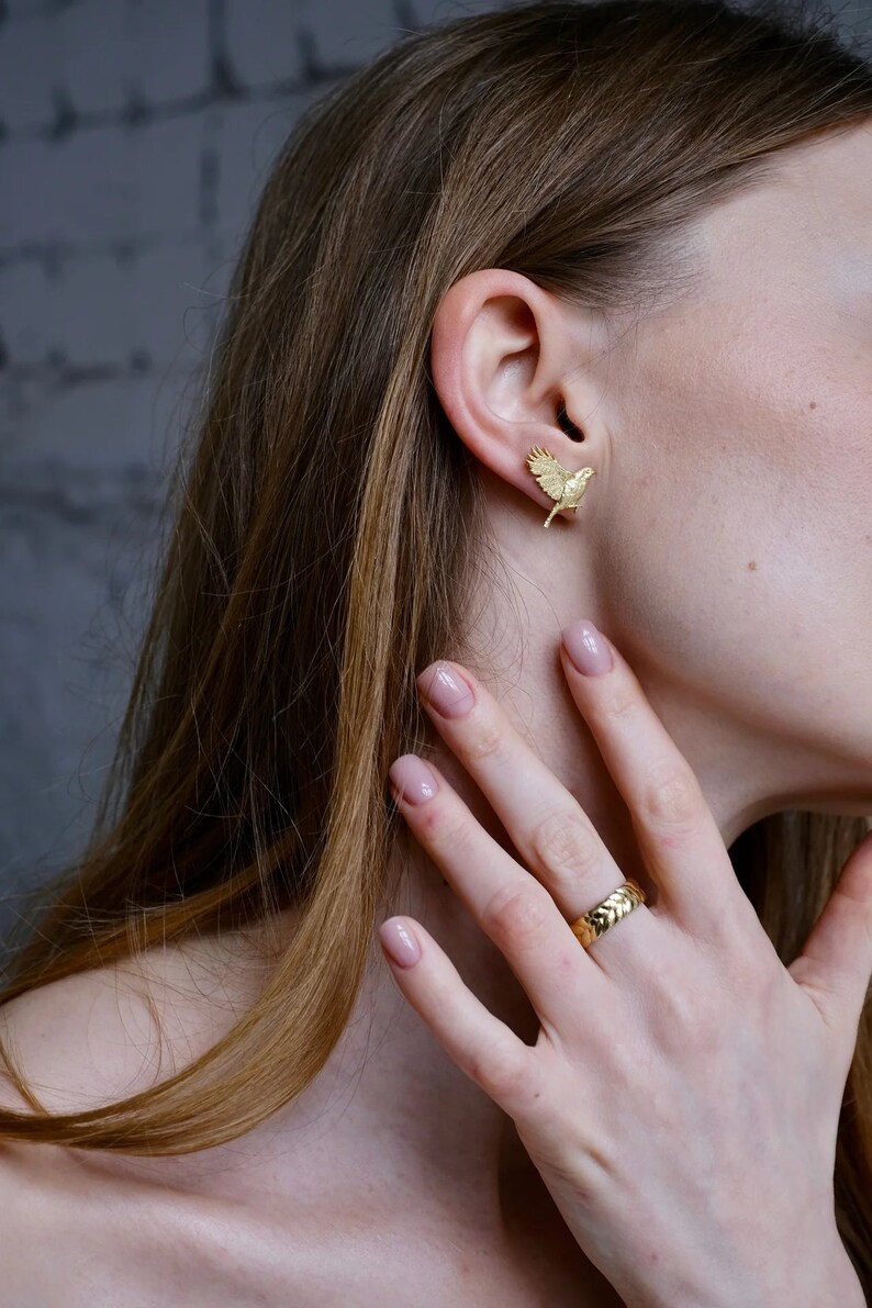May include: A gold bird-shaped earring with a textured surface. The earring is worn in the left ear of a person with long brown hair. The person is also wearing a gold ring on their right ring finger.