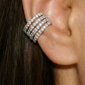 Puede incluir: Un brazalete de oreja brillante con cuatro filas de pequeñas piedras preciosas transparentes. La joya envuelve la oreja, creando un accesorio glamuroso y llamativo. El brazalete se coloca sobre cabello oscuro y piel.