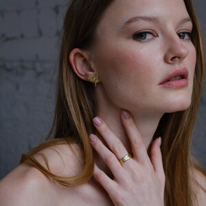 May include: A woman wearing a gold ring with a textured design on her finger. She is also wearing a pair of gold earrings with a leaf-like design.