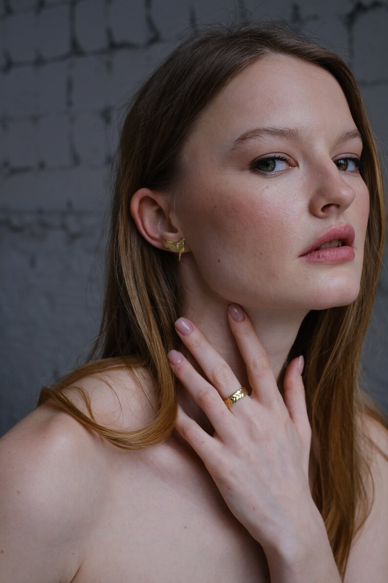 May include: A woman wearing a gold ring with a textured design on her finger. She is also wearing a pair of gold earrings with a leaf-like design.