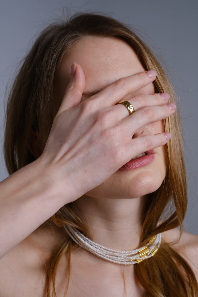 May include: A woman wearing a gold ring and a multi-strand pearl necklace with gold accents.