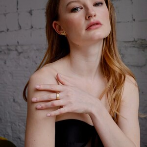 May include: A woman with long, wavy, light brown hair is wearing a black, sheer, strapless top. She is wearing a gold ring on her finger and gold earrings.