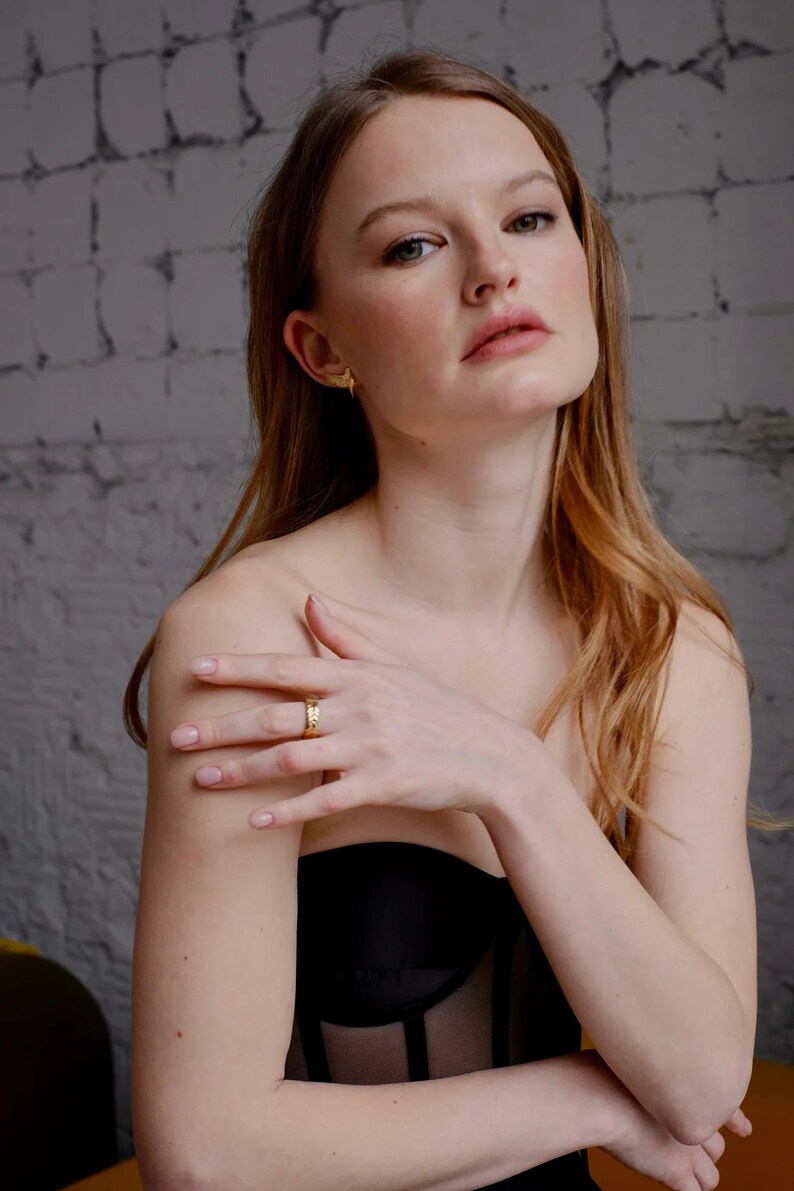 May include: A woman with long, wavy, light brown hair is wearing a black, sheer, strapless top. She is wearing a gold ring on her finger and gold earrings.