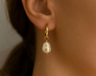 Puede incluir: Un pendiente de aro dorado con una perla colgante en forma de lágrima. El pendiente tiene un diseño sencillo y elegante, con una pequeña cuenta dorada en la parte inferior de la perla. El fondo está borroso.