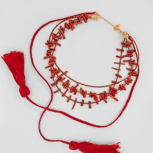 May include: A multi-strand necklace featuring red coral beads and red tassels. The necklace is crafted from braided red cord and has a gold clasp.